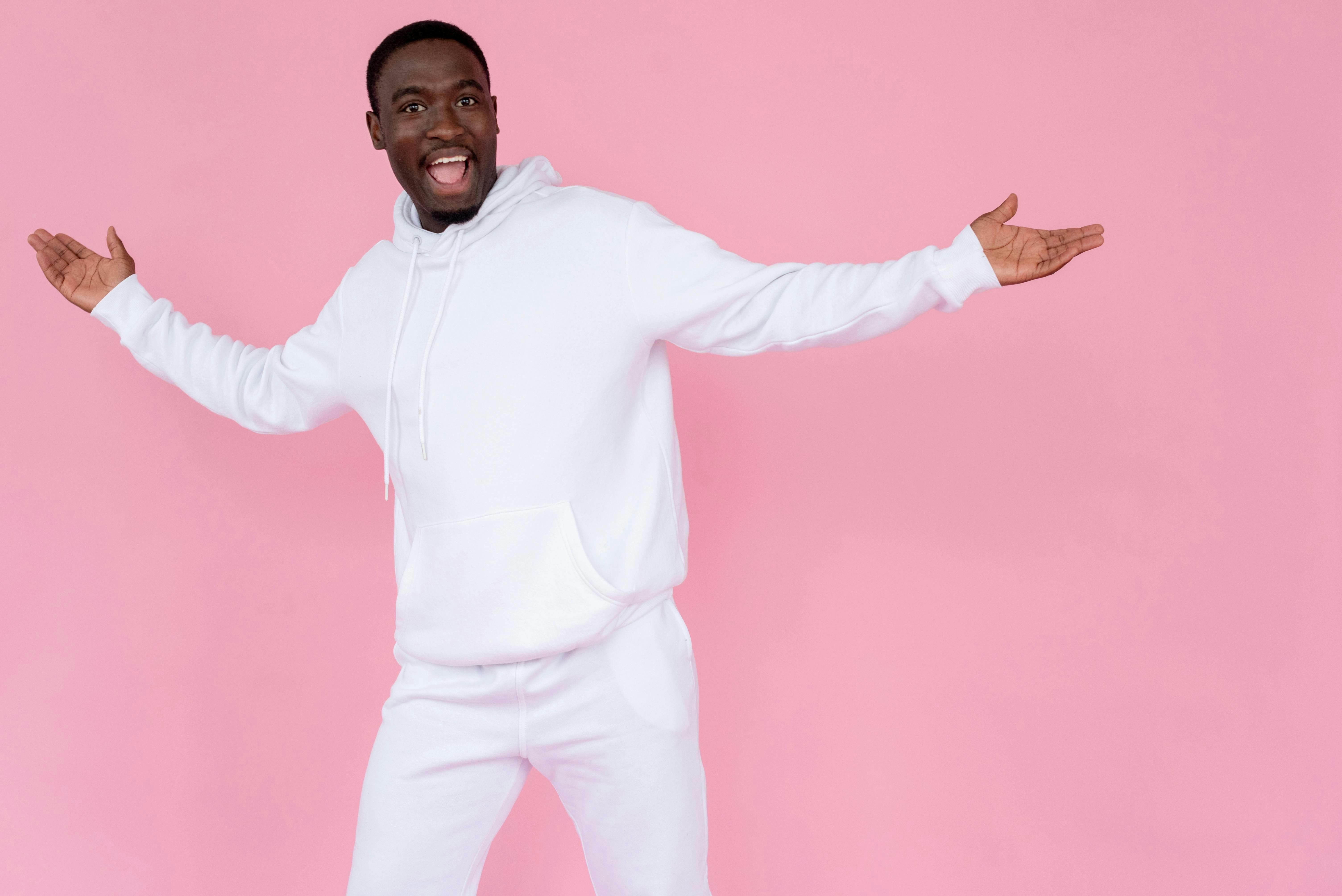 Man wearing all white sweat outfit smiling with hands out on pink background