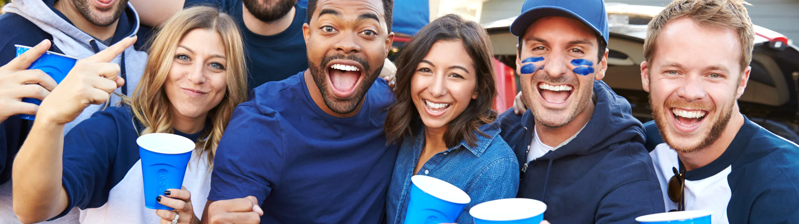 A group of smiling people wearing casual clothing holding blue cups, posing energetically outdoors