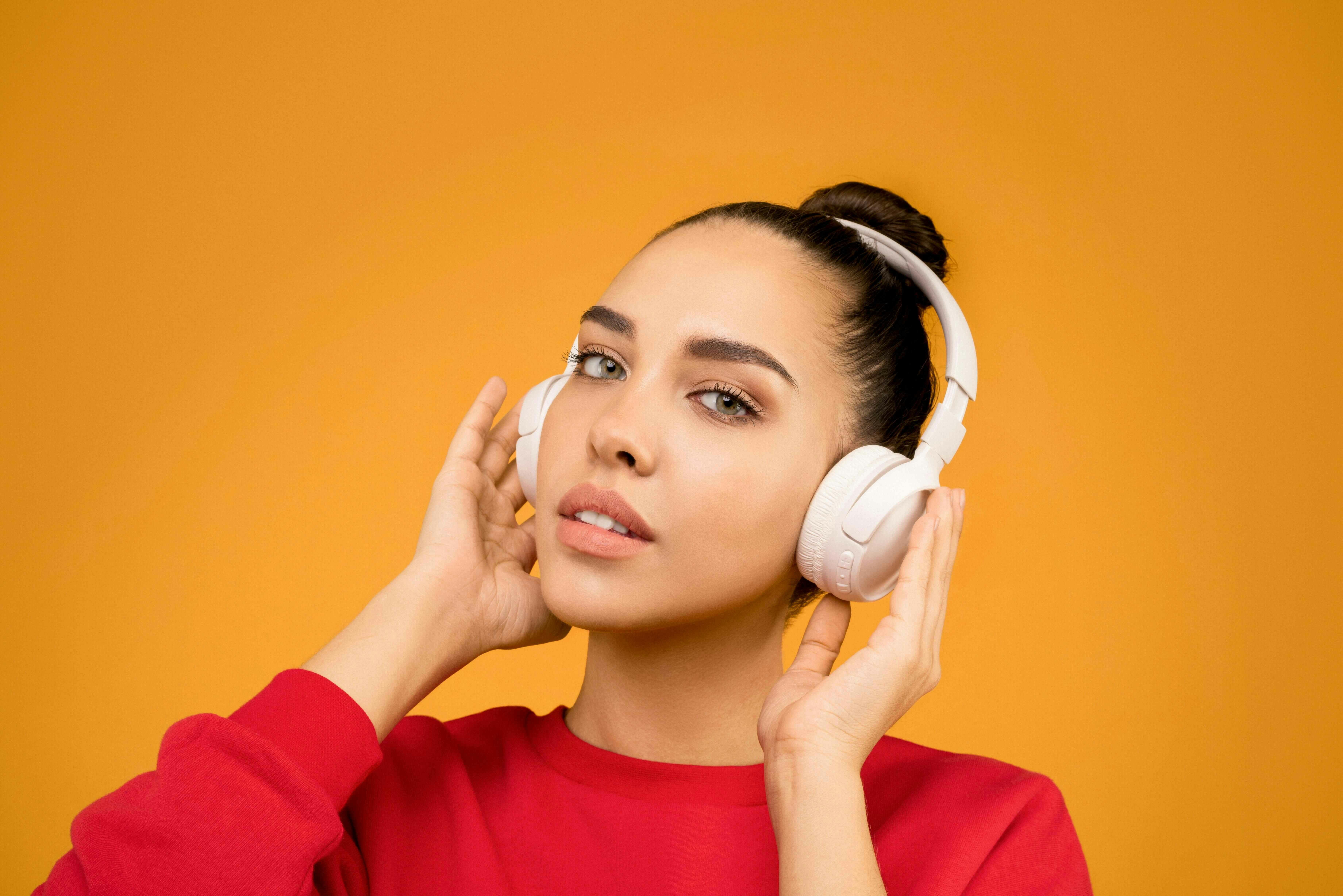 Woman with hair in a bun in a red sweatshirt holding over ear white headphones