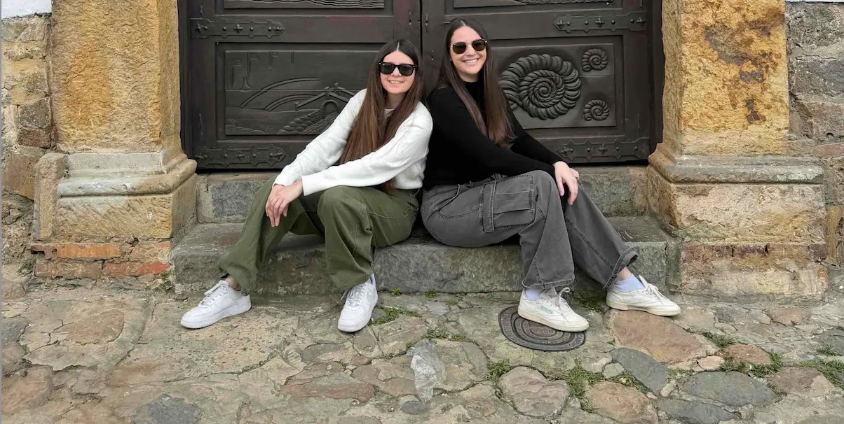Two women sitting back to back on the stone steps of a building, framed by a doubledoor entrance.