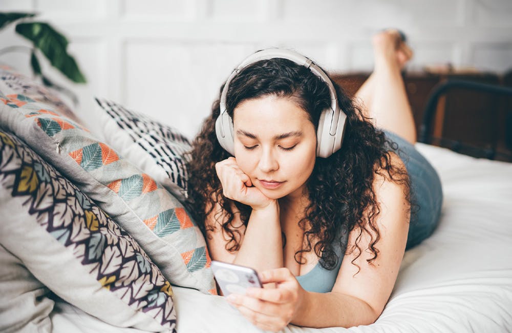 woman laying on bed holding cell phone and wearing headphones