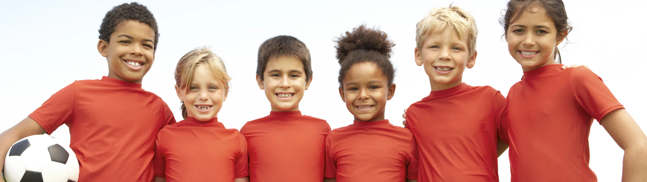 Six children standing together, smiling, and wearing red shirts. One child is holding a soccer ball.